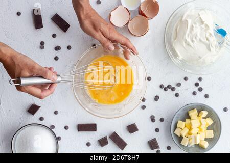Top view crop unrecognizable cook beating eggs in bowl placed on table with baking ingredients including butter sour cream and chocolate Stock Photo