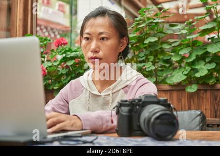 Concentrated Asian female journalist sitting at table with digital photo camera and using laptop while preparing new article Stock Photo