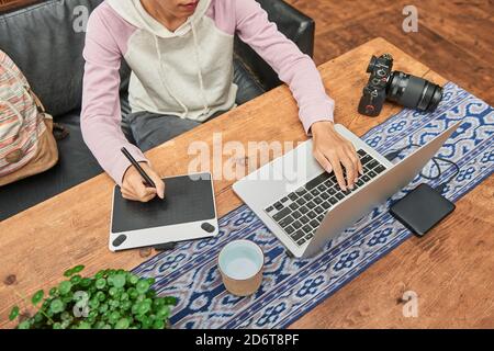 From above of crop anonymous female photographer using graphic tablet and laptop while sitting at table with photo camera and working on project Stock Photo