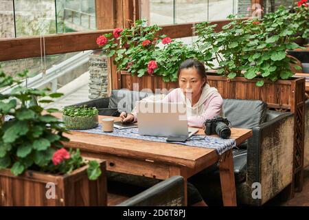 Concentrated Asian female journalist sitting at table with digital photo camera and using laptop while preparing new article Stock Photo