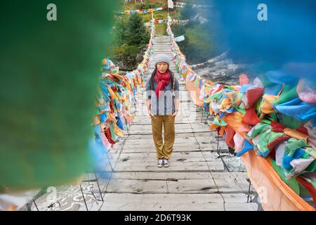 Young ethnic female tourist in warm clothing and hat looking at camera while crossing simple long narrow suspension wooden bridge decorated with brigh Stock Photo