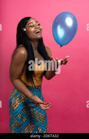 Happy young ethnic lady with long dark hair in stylish outfit standing and playing with balloon against pink background Stock Photo