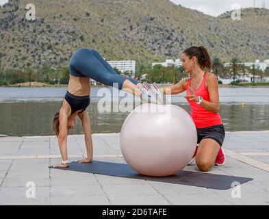 Enduring female athlete doing abdominal crunches while leaning on fitness ball during workout with help of professional trainer Stock Photo