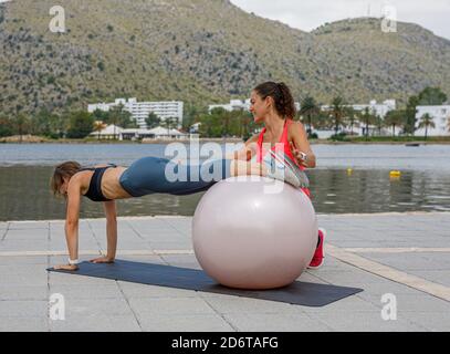 Enduring female athlete doing abdominal crunches while leaning on fitness ball during workout with help of professional trainer Stock Photo