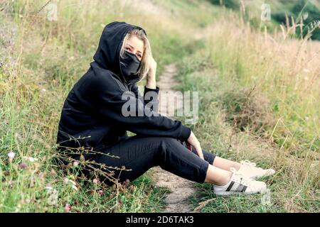 Beautiful blonde girl in a dark casual suit in a field with a black medical mask on her face. Stock Photo