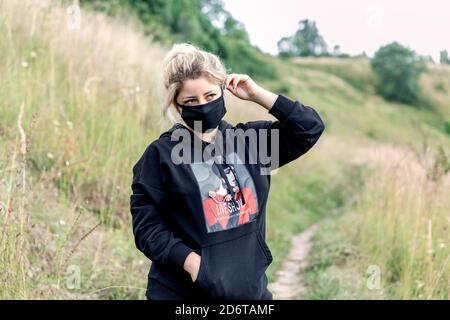 Beautiful blonde girl in a dark casual suit in a field with a black medical mask on her face. Stock Photo