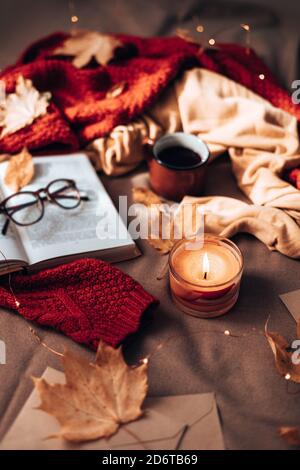 Burning candles, cup of hot tea, open book and glasses on plaid in bed. Autumn leaves and light garland decor on sofa. Autumn style. Hygge concept, vertical photo orientation Stock Photo