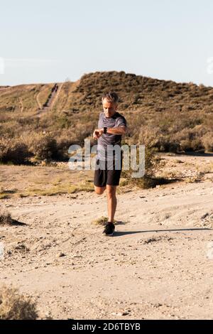 Full body of middle aged male in activewear checking time on smart watch while stretching leg during training in countryside Stock Photo