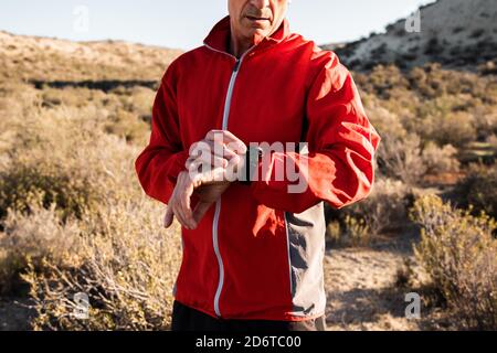 Concentrated middle aged male athlete in activewear checking time on smartwatch while standing in hilly terrain after workout Stock Photo