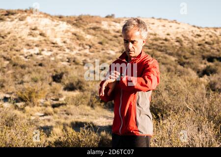 Concentrated middle aged male athlete in activewear checking time on smartwatch while standing in hilly terrain after workout Stock Photo