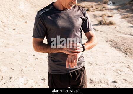 Concentrated middle aged male athlete in activewear checking time on smartwatch while standing in hilly terrain after workout Stock Photo