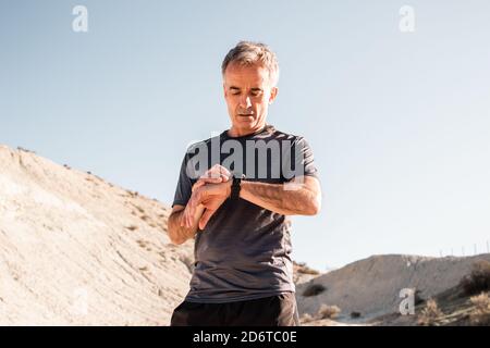 Concentrated middle aged male athlete in activewear checking time on smartwatch while standing in hilly terrain after workout Stock Photo