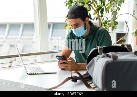 Smiling male wearing medical mask sitting at table with laptop and using cellphone Stock Photo