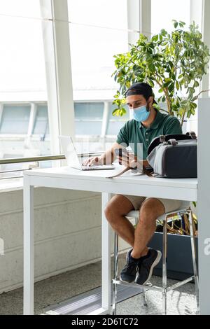 Male wearing medical mask sitting at table and using cellphone and laptop simultaneously Stock Photo