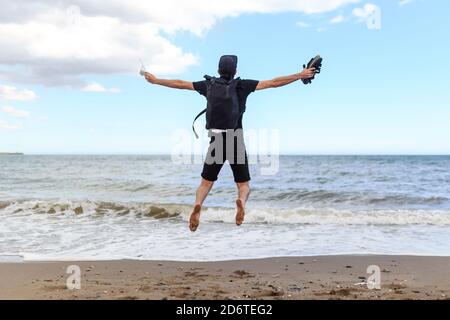 Back view of anonymous content male traveler with backpack jumping in air with raised arms over sandy shore near ocean under blue cloudy sky Stock Photo