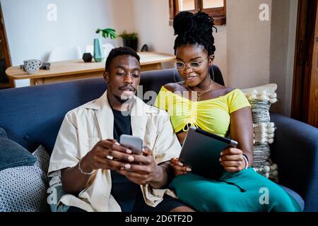 Content young African American lady using tablet while resting on sofa with boyfriend using smartphone at home Stock Photo