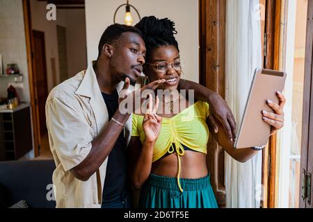 Positive young ethnic man cuddling cheerful girlfriend in crop top making two finger gesture while having video chat on tablet at home Stock Photo