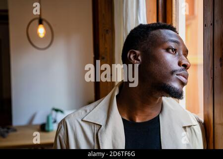 Pensive young African American guy in casual clothes looking out window while resting at home in daylight Stock Photo