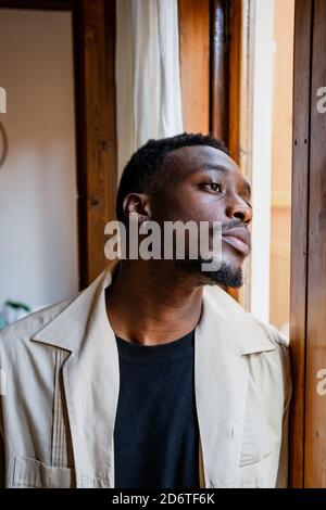 Pensive young African American guy in casual clothes looking out window while resting at home in daylight Stock Photo
