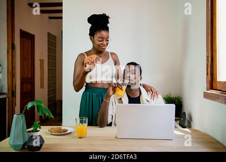 Concentrated black male freelancer working at laptop while thoughtful African American wife in crop top eating bun and looking away Stock Photo