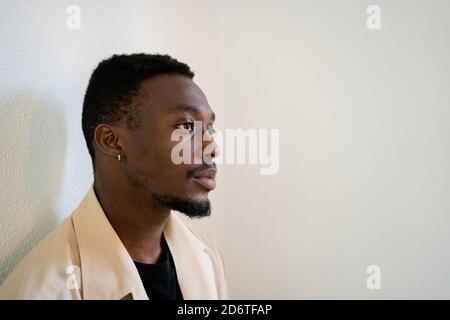 Pensive young African American male in light beige formal shirt thoughtfully looking away while standing near white wall Stock Photo