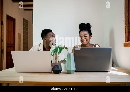Front view of positive young African American man and Woman in casual clothes and smiling while working together on laptops at home Stock Photo