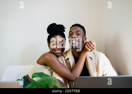 Positive young slim African American wife in casual clothing looking at camera with smile while embracing husband in bright room decorated with green Stock Photo
