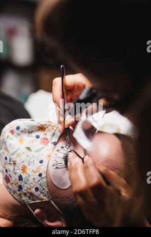 From above of crop anonymous master doing eyelash extension after mapping for unrecognizable female customer in face mask Stock Photo
