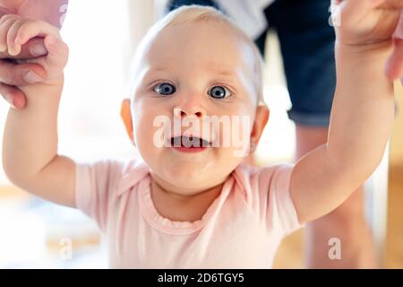 Cute baby with blond hair and light blue sky looking at camera with mouth opened while holding hands of faceless parent Stock Photo