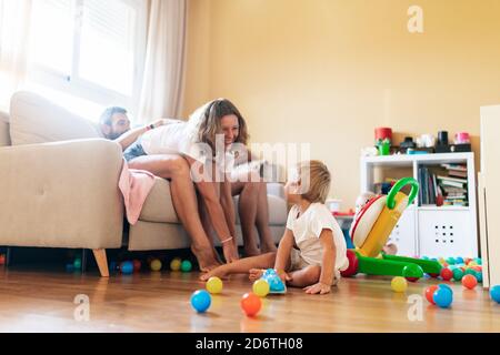 Full body of cheerful mother sitting on couch with husband and playing with baby in cozy living room Stock Photo