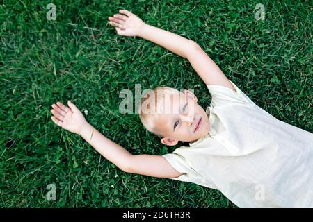 Overhead cheerful blond haired boy in white shirt looking at camera with smile while chilling on verdant grass with arms behind head Stock Photo