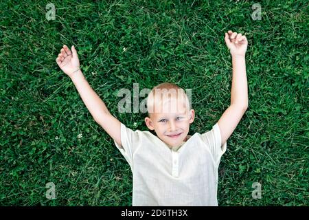 Overhead cheerful blond haired boy in white shirt looking at camera with smile while chilling on verdant grass with arms behind head Stock Photo