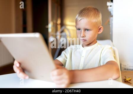 Side view of concentrated blond haired little boy in white shirt browsing internet on tablet in bright room in sunlight Stock Photo