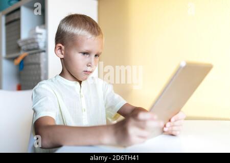 Side view of concentrated blond haired little boy in white shirt browsing internet on tablet in bright room in sunlight Stock Photo