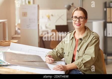 Portrait of smiling young woman drawing blueprints and plans while working at desk in engineers office, copy space Stock Photo