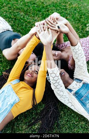 High angle of happy multiracial girlfriends lying together on grass and stacking hands while resting in park in summer Stock Photo