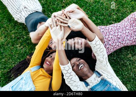 High angle of happy multiracial girlfriends lying together on grass and stacking hands while resting in park in summer Stock Photo