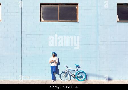 Full body of trendy bearded male in stylish outfit and sunglasses leaning on blue wall while relaxing on street near modern bicycle on sunny day Stock Photo
