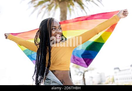 Low angle of proud African American female standing with fluttering LGBT rainbow flag on street and cheerfully looking away Stock Photo