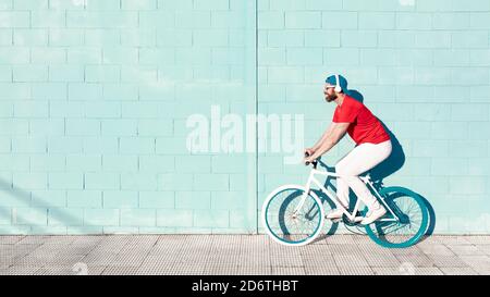 Side view of young active bearded male cyclist in stylish outfit listening to music in headphones while riding bike on street near blue stone building Stock Photo