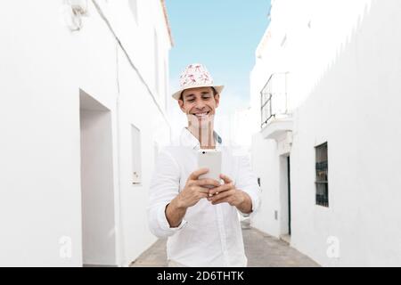 Young cheerful trendy male tourist in hat text messaging on cellphone between old houses in White Village Stock Photo