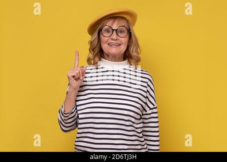 Smiling mature woman in yellow hat and glasses pointing finger up Stock Photo