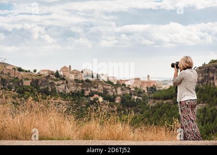 Side view of unrecognizable mature female photographer with professional photo camera taking pictures of old town Cuenca with medieval buildings on hi Stock Photo