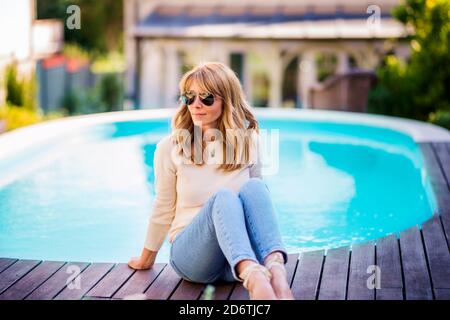Confident smiling woman wearing casual clothes while relaxing on the poolside at home. Stock Photo