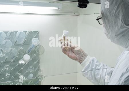 Scientist in protective white coat, mask and gloves analyzes a virus or bacteria sample in a laboratory with vials, glass flasks and chemicals Stock Photo
