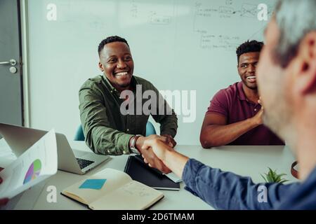 Smiling mixed race business man shaking hands with manager accepting project deal sitting in boardroom during team meeting  Stock Photo