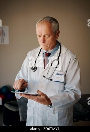 Handsome senior doctor with serious expression standing with labcoat and stethoscope using digital tablet Stock Photo