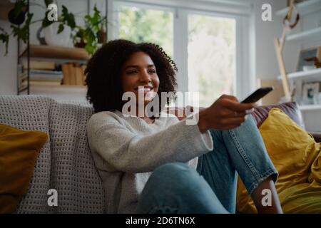 Portrait of cheerful woman sitting on couch at home watching tv and holding remote control Stock Photo