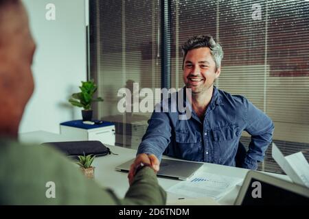 Caucasian boss shaking hands with ethnic colleague accepting business deal sitting in office  Stock Photo