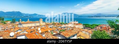 Panorama of Desenzano del Garda town with red tiled roof buildings, Garda Lake water, Monte Baldo mountain range, Sirmione peninsula, Lombardy, Northern Italy. Aerial panoramic view of Desenzano Stock Photo